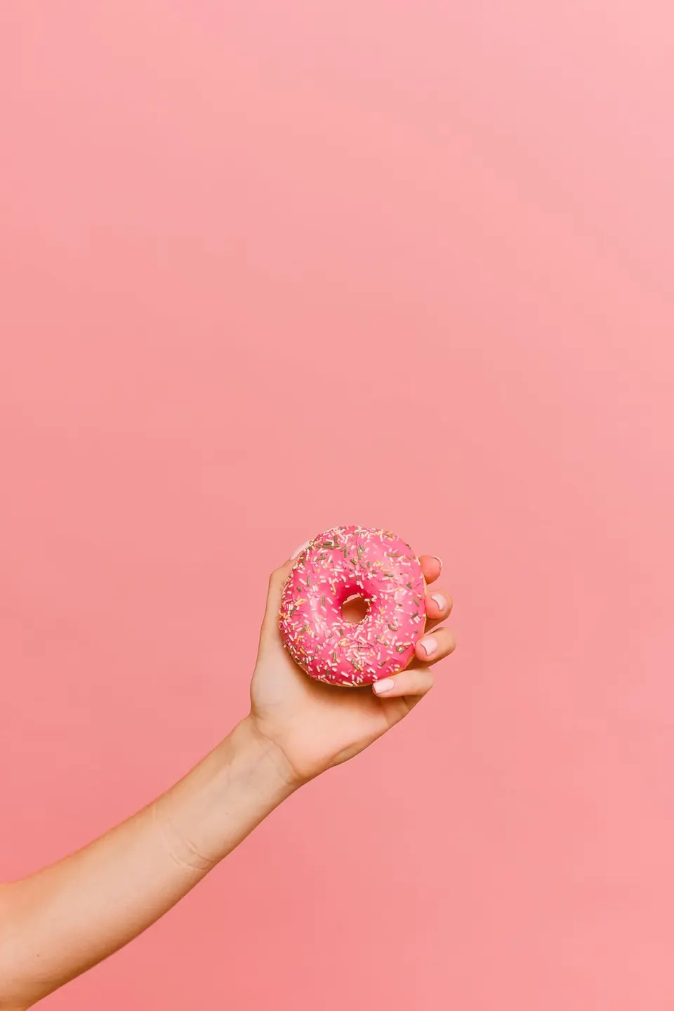 Mano sosteniendo una dona rosada con confites sobre fondo rosa